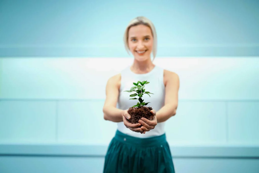 Frau hält eine kleine Pflanze mit Erde in beiden Händen und lächelt – Symbolbild für Nachhaltigkeit.